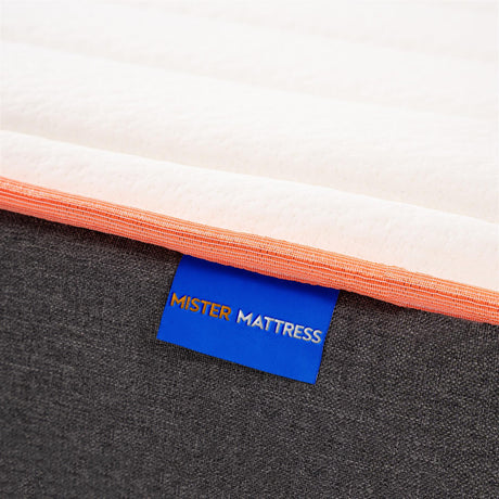 Close-up of a mattress corner showing a white top surface, an orange stitched edge, a dark gray fabric underside, and a blue tag with the brand name MISTER MATTRESS.