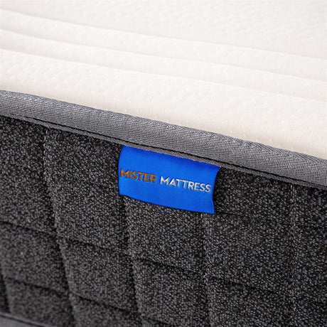 Close-up of a mattress corner showing a white quilted top surface, dark gray textured side panel, and a blue fabric label reading MISTER MATTRESS.