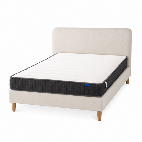 A mattress on a upholstered bed frame with a light beige headboard and legs, shown on a clean white background. The mattress has a white top surface and a dark charcoal quilted side panel with a small blue brand tag.