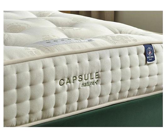 Close-up of a tufted mattress corner showing the side panel with the brand name CAPSULE natureX and a blue mattress tag, set against a neutral indoor background.