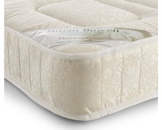 A close-up of a quilted mattress corner in a warm cream or off-white color, featuring a subtle floral damask pattern. A clear plastic window on the top shows the brand name 'Julian Bowen' and the model 'Platinum Book'.