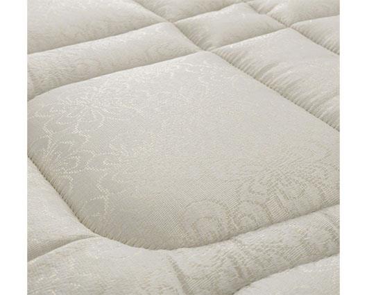 A close-up of a quilted mattress cover in a warm off-white/cream color, showing a subtle woven floral pattern on the central panel.