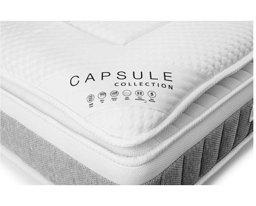 A close-up of a mattress corner showing a white quilted top layer with the text 'CAPSULE COLLECTION' and small certification icons, plus a gray ribbed side panel.